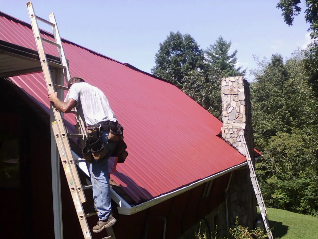 Roofer on a ladder beside a red metal roof and stone chimney — Soffit & Fascia Repair by Roofing Brothers in Barberton